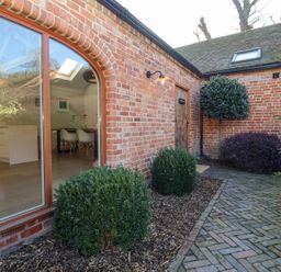 An outdoor brick walkway next to a building with large windows showing a kitchen and dining area at The Piggery in Ledbury near Welland