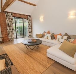 A living room with a white sectional sofa, cushions, round black coffee table, TV on a stand, and sliding glass door at The Piggery in Ledbury near Welland