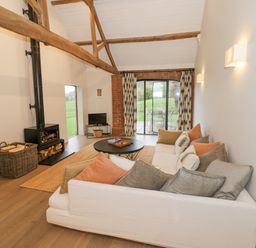 A living room with a white sofa and cushions a wood stove and basket with logs a round coffee table and glass doors at The Piggery in Ledbury near Welland