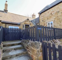 The stone exterior of a cottage with dark wooden fencing and stone steps leading to a door at Ash Cottage in Lucker near Warenford