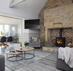 A living room with a stone fireplace, a wood burning stove, a wall-mounted TV, grey sofas, and a dining area with a round wooden table and chairs at Elm Cottage in Lucker near Warenford