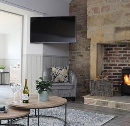A living room with a stone fireplace and wood burner a round coffee table with wine glasses a wine bottle and a plant an armchair a wall mounted television at Elm Cottage in Lucker near Warenford