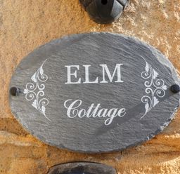 A grey slate oval house sign with the words Elm Cottage mounted on a stone wall at Elm Cottage in Lucker near Warenford