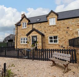 A stone house with black fencing and a wooden bench in the gravel front yard at Yew Cottage in Lucker near Warenford