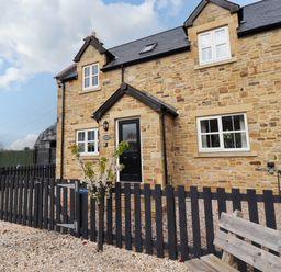 A stone house with a black front door and white windows behind a black wooden fence and gravel ground at Yew Cottage in Lucker near Warenford