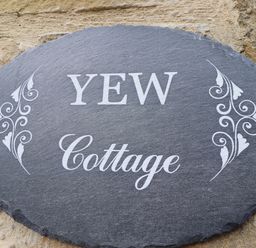 An oval slate house sign with white decorative patterns and the words Yew Cottage mounted on a stone wall at Yew Cottage in Lucker near Warenford