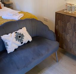 A room with a gray sofa with a patterned cushion a wooden cabinet with cups and glasses and a bed with towels and a yellow blanket at Red Squirrels Drey in Lucker near Belford