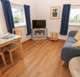 A living room with a blue sofa and yellow pillows a wooden dining table two windows with blue curtains and a television on a gray stand at Ramblers Rest in St Just