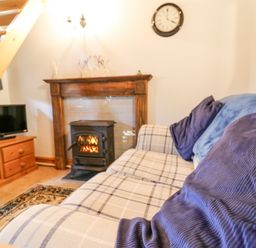 A living room with a sofa, cushions, a fireplace with a stove, a wooden TV stand, and a staircase at Ty Bach Twt in Synod Inn near New Quay
