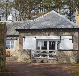 A small stone building with white wooden shutters and a gate surrounded by trees at Harness Lodge in Seahouses