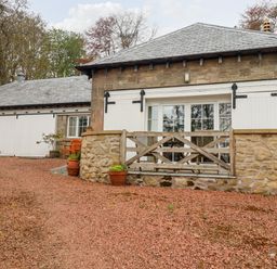 A stone building with white wooden doors and windows and a gravel driveway with potted plants at Harness Lodge in Seahouses