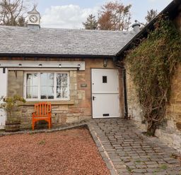 An exterior view of a stone building with a white door, a window, an orange wooden chair, and a small potted plant at Harness Lodge in Seahouses