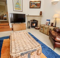 A living room with a wicker coffee table a leather armchair a wooden chest a fireplace with a wood stove and a TV on a wooden stand at Harness Lodge in Seahouses