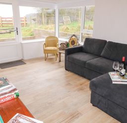 A sitting room with a black sofa a wooden chair a small table with wine bottle books and glasses and large windows overlooking a garden at The Poets Cottage in Portglenone