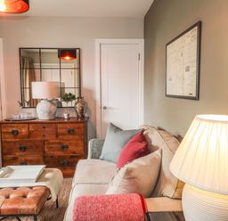 A living room with a beige sofa and colorful cushions a wooden chest of drawers with a lamp and decorative items a leather ottoman and a lamp at The Pottery in Newport Pembrokeshire