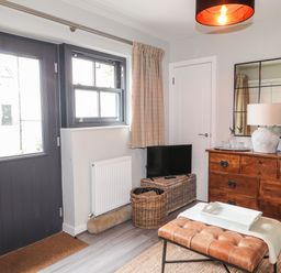 A living room with a dark door and window curtains a wooden dresser with a lamp and mirror and a leather bench at The Pottery in Newport Pembrokeshire