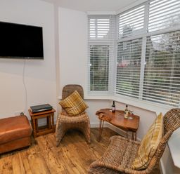 A corner seating area with two wicker chairs and plaid cushions a wooden table with bottles and glasses a wall-mounted television and a leather ottoman at Bronwen House in Llanbedr
