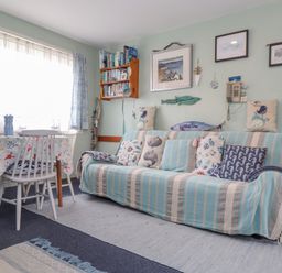A living room with a sofa covered in striped fabric and sea-themed pillows beside a dining table with chairs and a patterned tablecloth at Villa 29 in Camelford