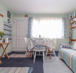 A dining area with a table and chairs by a window next to a sofa with cushions and a wooden side table with plants at Villa 29 in Camelford
