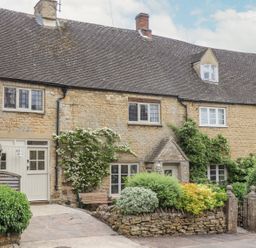 A stone cottage with a tiled roof and climbing plants on the walls surrounded by bushes and a small garden at Lynton Cottage in Enstone
