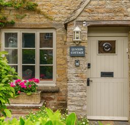 A stone cottage exterior with a beige door and window with flowers in front at Lynton Cottage Enstone