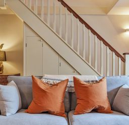 A sofa with orange and light grey cushions in front of a staircase with a wooden handrail at Lynton Cottage in Enstone