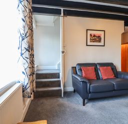 A room with a blue leather sofa with two red cushions a framed picture on the wall stairs with carpet and a window with patterned curtains at Ynys in Dyffryn Ardudwy