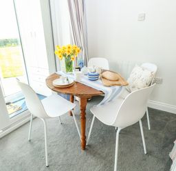 A dining area with a round wooden table set with dishware yellow flowers and bread surrounded by white chairs and a bench at Gull Cottage in Kessingland