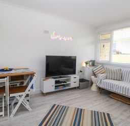 A living room with a TV on a white stand a wooden table with chairs and a striped sofa at Dollys Lookout in Kessingland