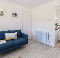 A small living room with a blue sofa and framed pictures on white walls next to a kitchen at Dollys Lookout in Kessingland