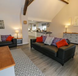 A living room with two black sofas with pillows a wooden table a sideboard and an open kitchen area at Bodrual Cottage in Rhosbodrual near Caernarfon