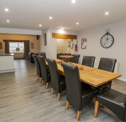 A dining area with a long wooden table and eight black chairs near a kitchen and living room at Bodrual Cottage in Rhosbodrual near Caernarfon