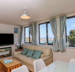 A living room with a beige sofa and blue cushions near windows with blue curtains and a wall mounted TV at Beachcomber in Porthcurno near Sennen