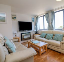 A living room with two beige sofas and blue cushions with a wooden coffee table and a wall-mounted TV at Beachcomber in Porthcurno near Sennen