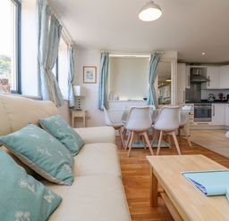 A living room with a cream sofa and turquoise cushions a dining table with white chairs and an open kitchen in the background at Beachcomber in Porthcurno near Sennen
