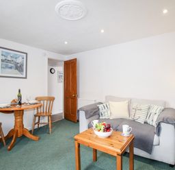 A living room with a sofa and cushions a coffee table with fruit a dining table with chairs and a bottle of wine at Angel Cottage in Weymouth