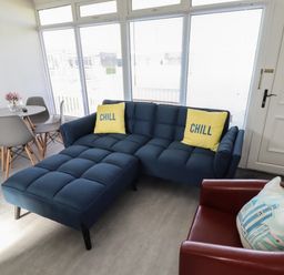 A living room with a blue sectional sofa with yellow pillows a round table with four chairs a red armchair and a wall clock at 23 Cherry Park in Chapel St Leonards