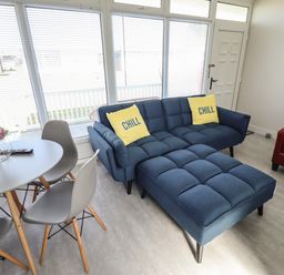 A living room with a blue sectional sofa with yellow chill pillows a round dining table with gray chairs and large windows at 23 Cherry Park in Chapel St Leonards