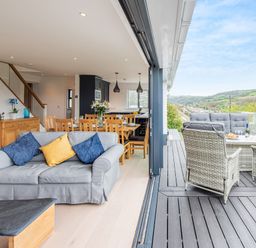 An open living and dining area with a gray sofa and wooden dining table next to an outdoor deck with wicker furniture and a view of green hills at Beach View in Combe Martin