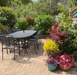 A patio with a black metal table and chairs surrounded by plants and flowers at Woodbine Cottage in Mylor Bridge Nr Falmouth
