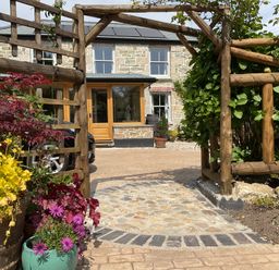 A stone house with wooden doors viewed through a wooden garden arch with potted plants at Woodbine Cottage in Mylor Bridge Nr Falmouth