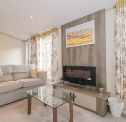 A living room with a beige sofa yellow pillows glass coffee table and wall-mounted television at Gwydn Lodge in Downton Hampshire