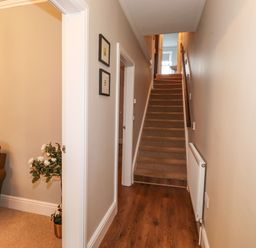 A hallway with wooden flooring leading to carpeted stairs and a partially visible room with a brown sofa and a potted plant at Snowflake Cottage in Windermere