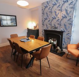 A dining room with a wooden table six brown chairs a black fireplace with floral decorations an orange armchair and blue patterned wallpaper at Snowflake Cottage in Windermere