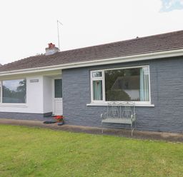A single story house with white and gray exterior walls a green metal bench on the front patio and a grass lawn at Glynderi in Cilgerran