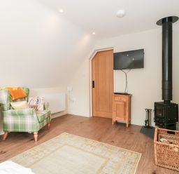 A room with a plaid armchair next to a wooden dresser a small wooden cabinet under a wall-mounted TV a wood-burning stove and a wicker basket with firewood at The Attic at Redmond Bottom in Wookey