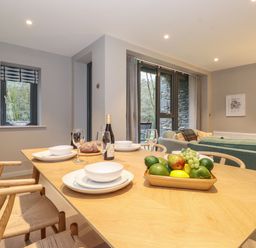 A dining table with plates glasses a bottle and fruit bowl next to a living area with sofas and large windows at Bruach in Backbarrow