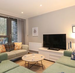 A living room with green and beige sofas around a round wooden coffee table and a TV on a white stand at Bruach in Backbarrow