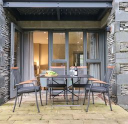 An outdoor patio with a table and four chairs in front of stone walls and glass doors at Bruach in Backbarrow
