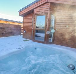 Outdoor hot tub on a snowy patio next to a wooden cabin with a glass door at Pegasus in Felmoor Park